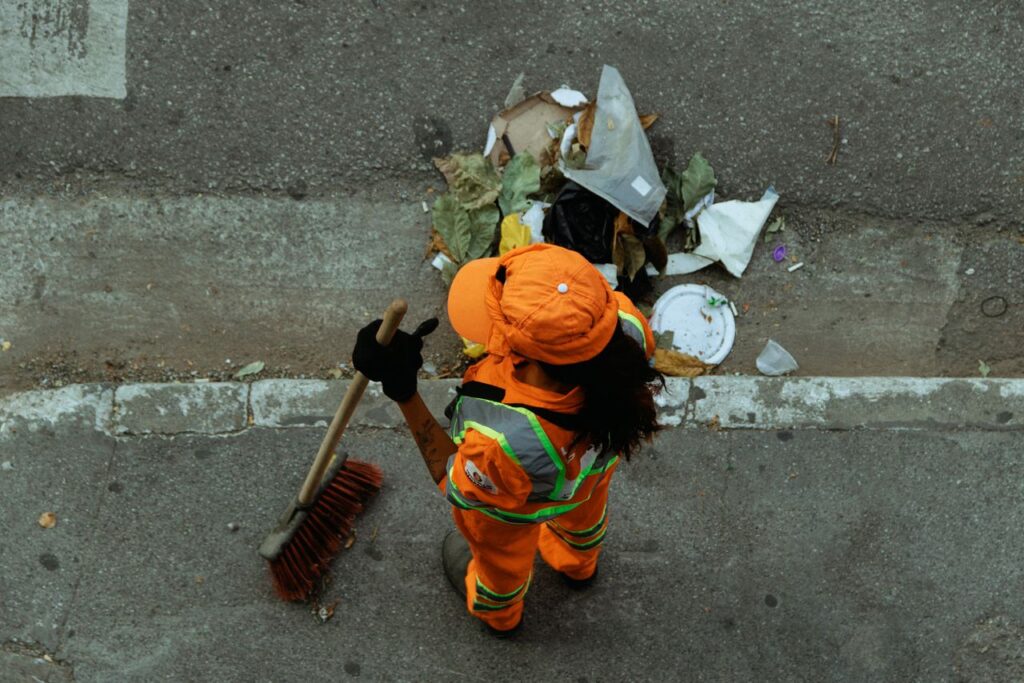 Top view of a street cleaner wearing orange uniform sweeping debris on a city sidewalk.